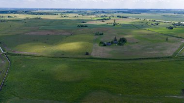 Uçsuz bucaksız kırsal bir arazide yeşil tarlalar ve dağınık ağaçlar bulutlu bir gökyüzünün altında..