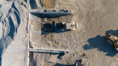 İnşaat sahasında çalışan ağır makinelerin havadan görüntüsü. Etrafı çakıl ve toprakla çevrili. Sahne, endüstriyel faaliyetleri vurgulayan bir yükleyici ve duvar yapısını gösteriyor..