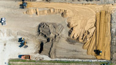 İçinde buldozerler ve kazıcılar, hareket eden ve kum ve kiri şekillendiren ağır makineler bulunan bir inşaat alanının havadan görüntüsü. Yerde çeşitli kum ve toprak yığınları görülebilir..