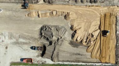 Kamyonlar ve kazıcılar da dahil olmak üzere araç ve ekipmanların bulunduğu bir inşaat alanının havadan görüntüsü bir otoparkın yanındaki toprak alanda çalışıyor..