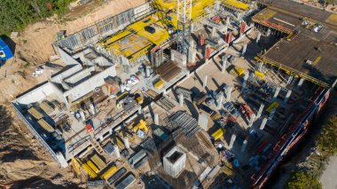 Vinçleri, iskelesi ve inşaat malzemeleri olan bir inşaat alanının havadan görüntüsü. İşçiler mevcut ve beton sütunlar ve kirişler gibi çeşitli inşaat elementleri görülebilir durumda..