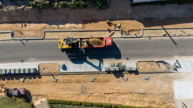 Sarı bir kazıcının kırmızı bir kamyona toprak yüklemesi de dahil olmak üzere ağır makineleri gösteren bir inşaat alanının havadan görüntüsü. Yol asfalt döşeniyor, işçiler ve malzemeler kenarda görünüyor..