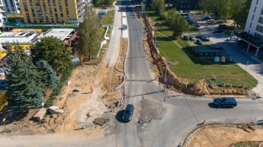 Şehir merkezindeki bir yol inşaatı alanının hava görüntüsü, kısmen tamamlanmış yollar ve inşaat ekipmanları arasında gezinen araçları gösteriyor. Arka planda binalar ve ağaçlar görünür..