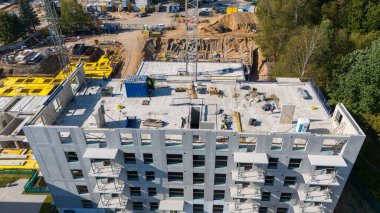 Kısmen inşa edilmiş çok katlı bir inşaat alanının havadan görüntüsü. Arazi ağaçlarla çevrili ve inşaat ekipmanları görünür durumda. Çatıda işçiler var..