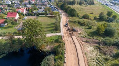 Yerleşim alanı yakınlarında inşaat halindeki bir kırsal yolun havadan görünüşü ve inşaat alanında makineler çalışıyor. Manzara yeşillik, gölet ve yakınlardaki evleri içerir..