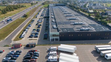 Çatıda güneş panelleri olan, araba ve kamyonlarla dolu otoparklarla çevrili büyük bir endüstriyel deponun havadan görüntüsü. Yakınlarda bir otoyol ve yerleşim alanı görülebilir..