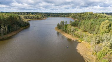 Bulutlu bir gökyüzünün altında yemyeşil ormanlarla çevrili sakin bir nehir manzarası. Suda iki küçük tekne görünüyor, bu da balık tutma veya boş zaman aktivitelerini gösteriyor..