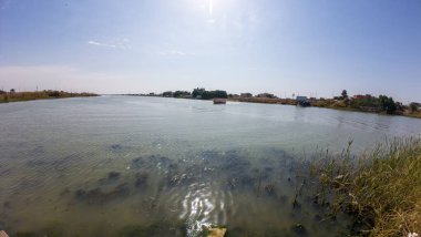 Sakin bir nehir manzarası. Etrafı bitki örtüsü ve açık mavi gökyüzünün altındaki uzak binalarla çevrili..