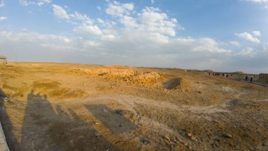 Parçalı bulutlu bir gökyüzünün altında dağılmış kayalarla çorak bir çöl manzarası..