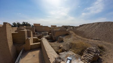 Parlak bir gökyüzü altında antik kalıntılar, taş duvarlar ve yollar, bir çöl manzarasıyla çevrili. Tarihi eser, eski yapıların kalıntıları ve ağaçların uzak bir görüntüsüyle ortaya çıkıyor..