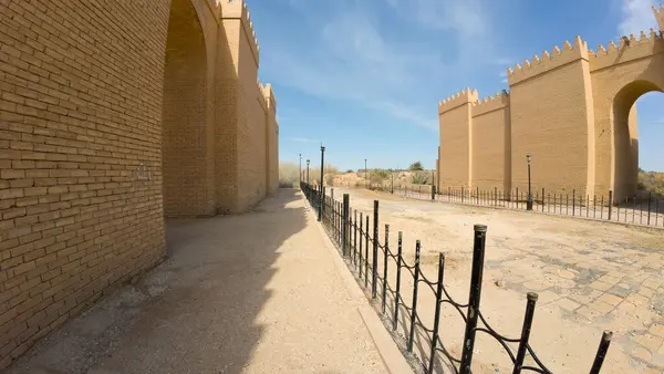Açık mavi gökyüzü ile antik şehir duvarlarının panoramik bir görüntüsü. Duvarlar sarı tuğlalardan yapılmış, tarihi mimari sergileniyor. Duvarların yanında demir bir çitle çevrili bir patika var..
