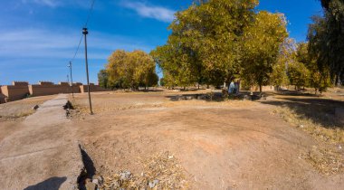 Açık mavi gökyüzünün altında sarı yapraklı büyük ağaçların yer aldığı kuru bir manzaranın panoramik bir görüntüsü. Toprak çoğunlukla bazı çimen parçaları ile çıplak ve taş yollar var.