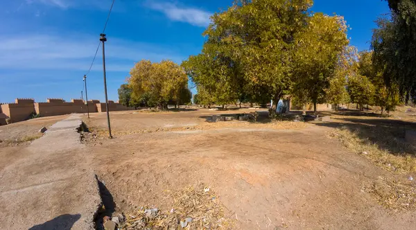 Açık mavi gökyüzünün altında sarı yapraklı büyük ağaçların yer aldığı kuru bir manzaranın panoramik bir görüntüsü. Toprak çoğunlukla bazı çimen parçaları ile çıplak ve taş yollar var.