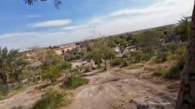 Çöl manzarası seyrek bitki örtüsü, ağaçlar ve uzak bir yol. Gökyüzü parçalı bulutlu ve arazi yeşillik parçalarıyla kumlu..