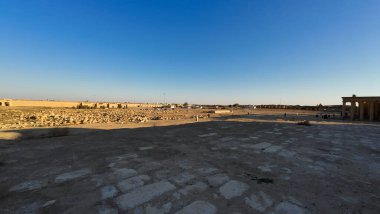 Temiz mavi gökyüzünün altındaki antik bir arkeolojik alanın geniş açılı görüntüsü, çöl manzarasında dağınık harabeler ve taş yapılar..