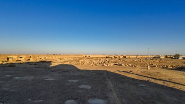 Ufukta engin mavi gökyüzü altında engin bir çöl manzarası, dağınık harabeler ve uzak yapılar görünüyor..
