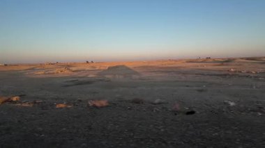 Açık mavi gökyüzünün altında geniş bir çöl manzarası, ufukta birkaç uzak ağaç ve yapılar var..