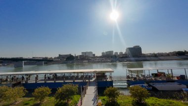 Arka planda rıhtım ve şehir silueti olan güneşli bir nehir kenarı sahnesi..