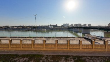 Tuğladan bir duvarı, park edilmiş araçları ve parlak bir güneşin altındaki uzak şehir manzarası olan sakin bir nehir manzarası..