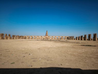 Arka planda belirgin bir kemer yolu ve yüksek bir yapıya sahip antik kalıntıların yer aldığı uçsuz bucaksız, çorak bir manzara. Gökyüzü açık ve mavi, sahnenin ıssızlığını vurguluyor..