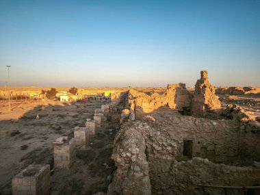 Gün batımında çöl manzarasında eski bir taş yapının kalıntıları, dağınık bitki örtüsü ve uzak modern binalar. Sahne bir tarih ve izolasyon hissi uyandırıyor..