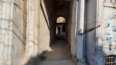 Grafiti ve enkazla yıkılan eski binalar arasında dar, yıpranmış bir ara sokak. Güneş ışığı duvarlara gölge düşürür, gizemli ve terk edilmiş bir atmosfer yaratır..