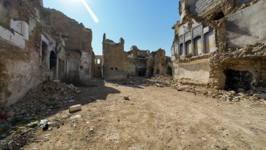 Terkedilmiş, savaştan zarar görmüş bir cadde. Paramparça olmuş binalar ve enkazlar berrak mavi gökyüzü altında..