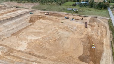 Buldozerler ve kamyonlar da dahil olmak üzere, büyük ve temiz bir arazi üzerinde çalışan ağır makineli bir inşaat alanının havadan görüntüsü. Manzara çoğunlukla çorak ve arka planda biraz yeşillik var..