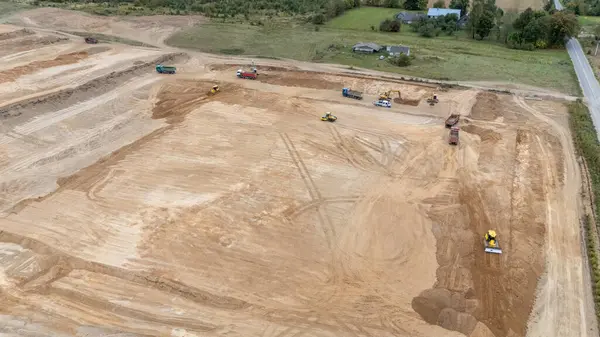 Buldozerler ve kamyonlar da dahil olmak üzere, büyük ve temiz bir arazi üzerinde çalışan ağır makineli bir inşaat alanının havadan görüntüsü. Manzara çoğunlukla çorak ve arka planda biraz yeşillik var..