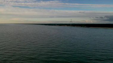 Ufukta bulutlu bir gökyüzünün altında rüzgâr türbini olan sakin bir kıyı manzarası..