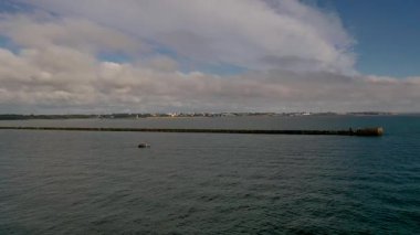 Parçalı bulutlu bir gökyüzünün altında sakin denize uzanan uzun dalgalı bir kıyı sahnesi..