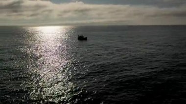 Bulutlu bir gökyüzünün altında parıldayan suya karşı silueti olan sakin bir okyanus sahnesi..