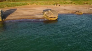 Kıyı manzarası, kumlu bir sahilde büyük beton bir sığınak, kayalık kayalıklar ve yeşil bitki örtüsüyle çevrili, açık bir gökyüzünün altında kıyıya vuran hafif dalgalar..
