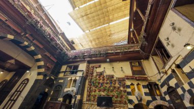 Asılı bitkiler ve karmaşık ahşap işçiliğiyle süslenmiş yüksek tavanlı geleneksel bir Orta Doğu avlusu sıcak ve davetkar bir atmosfer yaratıyor. Şam 'ın eski kasabasında