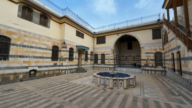 Geleneksel bir Orta Doğu avlusu. Çizgili taş duvarları, kemerli pencereleri ve merkezi bir çeşmesi var. Mimaride karmaşık desenler ve sakin bir atmosfer var. Şam 'ın eski kasabasında