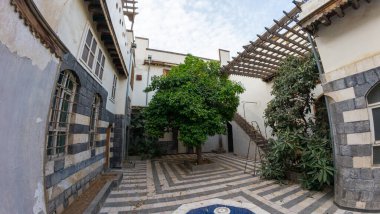 Merkezi bir ağacı olan sakin bir avlu. Parçalı bulutlu bir gökyüzünün altında, çizgili taş duvarlar ve ahşap sarkıtlar içeren geleneksel mimari yapılarla çevrili. Şam 'ın eski kasabasında