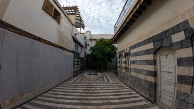Çizgili taş duvarları, küçük merkezi bir çeşmesi ve bir ağacı olan dar bir avlu. Gökyüzü parçalı bulutlu ve mimaride geleneksel Orta Doğu tasarımı yer alıyor. Şam 'ın eski kasabasında
