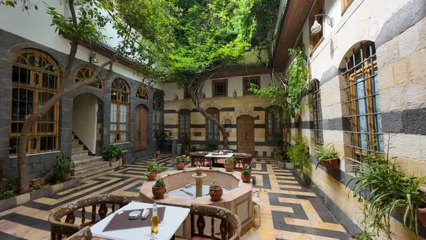 Merkezi bir çeşme, kemerli pencereler ve yemyeşil yeşillik içeren geleneksel Orta Doğu mimarisine sahip sakin bir avlu. İçkili masalar avluda hazır. Şam 'ın eski kasabasında