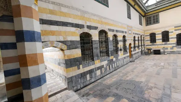 Çizgili taş duvarları ve kavisli pencereleri olan tarihi bir avlu, tipik bir Orta Doğu mimarisi olan sarı, mavi ve gri taşlardan oluşan bir karışım içeriyor. Şam 'ın eski kasabasında