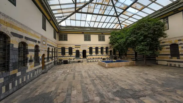 Cam çatılı boş bir avlu, içinde sarı ve gri çizgili taş duvarlar, ortada küçük bir çeşme ve birkaç ağaç var. Şam 'ın eski kasabasında