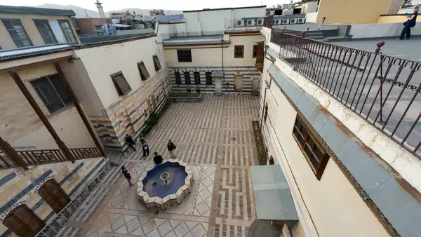 Eski binalarla çevrili, desenli taş döşemeli tarihi bir avlu manzarası. İnsanlar bir çeşmenin etrafında toplanıyor. Şam 'ın eski kasabasında