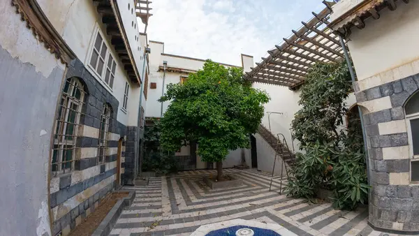 Merkezi bir ağacı olan sakin bir avlu. Parçalı bulutlu bir gökyüzünün altında, çizgili taş duvarlar ve ahşap sarkıtlar içeren geleneksel mimari yapılarla çevrili. Şam 'ın eski kasabasında