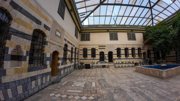 Geleneksel bir Orta Doğu avlusu. Çizgili taş duvarları, kemerli pencereleri ve cam çatının altında merkezi bir çeşmesi var. Şam 'ın eski kasabasında