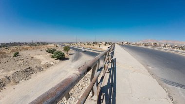 Metal parmaklıkları, çorak manzarası ve açık mavi gökyüzünün altındaki uzak binaları olan ıssız bir otoyolun geniş açılı görüntüsü. Suriye 'nin Şam Jobar ilçesinde