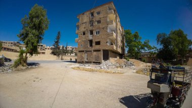 Önünde kırık camlar ve enkaz olan, berrak mavi bir gökyüzünün altında harap bir apartman. Yakınlarda sepetli bir ağaç ve motosiklet görünüyor. Bu da şehir ortamını işaret ediyor..