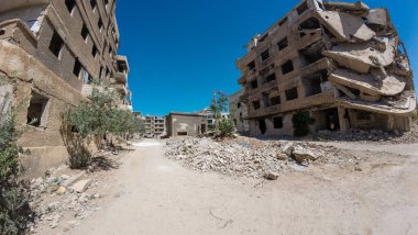 Hasarlı ve terk edilmiş binaların, yere saçılmış molozların ve açık mavi gökyüzünün bulunduğu ıssız bir şehir manzarası. Sahne bir yıkım ve ihmalkarlık hissi uyandırıyor..