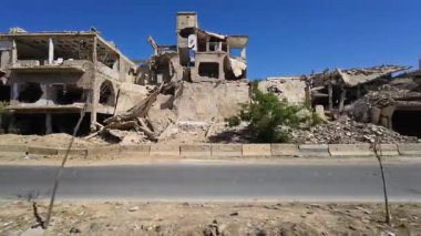 Paramparça olmuş, kısmen çökmüş tuğla ve molozlarla kaplı binalar, açık mavi gökyüzüne karşı kurulmuş. Arabadan sürerken çekilmiş. Suriye 'nin Şam Jobar ilçesinde