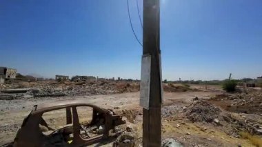 Açık mavi gökyüzünün altında hareket eden bir arabanın enkazı ve molozlarıyla çorak bir arazi. Suriye 'nin Şam Jobar ilçesinde