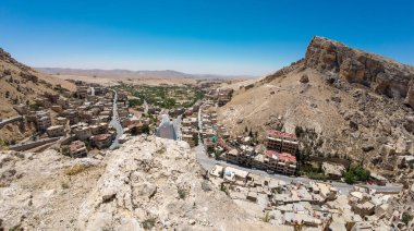 Açık mavi bir gökyüzünün altında kayalık tepelerin arasına kurulmuş bir Orta Doğu kasabasının panoramik görüntüsü. Maaloula Suriye.