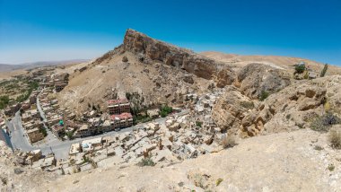 Açık mavi gökyüzünün altında dik bir tepeye kurulmuş kayalık bir çöl köyünün panoramik görüntüsü. Maaloula Suriye.